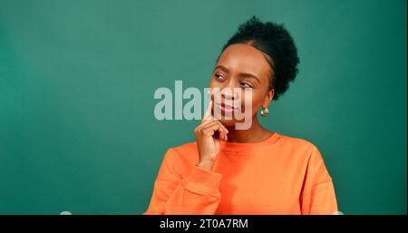 Serious young woman taps chin while thinking, contemplating, green studio Stock Photo