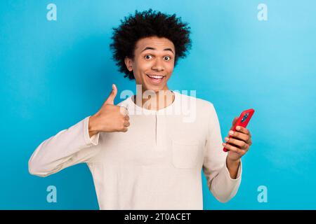 Photo portrait of nice young guy showing thumb up hold telephone wear trendy white pajama outfit isolated on blue color background Stock Photo