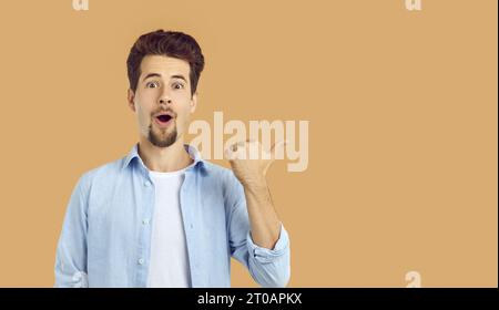 Happy, surprised, funny, handsome, young man with beard and mustache, dressed in casual white T-shirt and blue shirt, points with his thumb to the Stock Photo