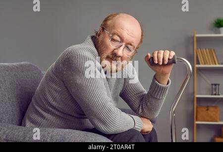 Sad, thoughtful, red-haired, elderly man with beard and glasses is sitting in gray armchair at home. Stock Photo