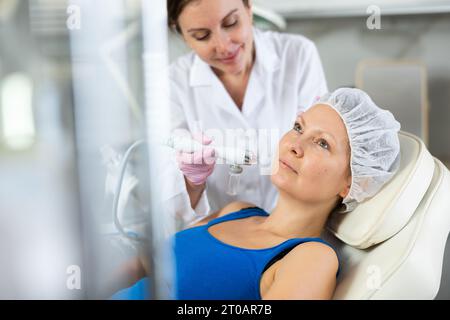 Beautiful female doctor making facial gas-liquid oxygen water epidermal peeling using macromolecule atomizing pen for adult woman at beauty salon.  Stock Photo