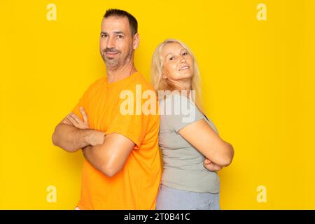 Happy loving couple. Studio shot of a beautiful couple in their 40s standing back to back and smiling, isolated over yellow background. Stock Photo