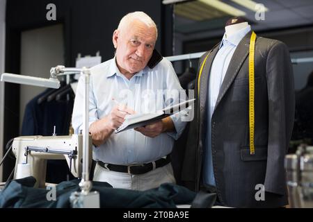 Professional tailor communicates with the client on mobile phone and takes notes in notebook Stock Photo