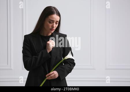 Sad woman with calla lily flower near white wall, space for text. Funeral ceremony Stock Photo