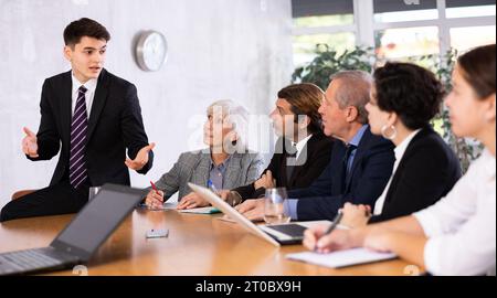 Young manager sitting on table during meeting with colleagues Stock Photo