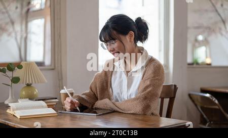 A happy and pretty Asian female college student working remotely at a cafe, using her digital tablet at a table indoors. People and technology concept Stock Photo