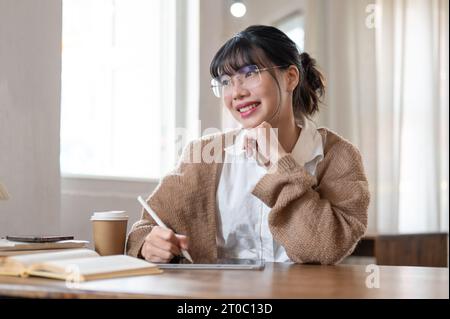 A beautiful and inspired young Asian woman in comfy clothes working remotely at a coffee shop, using her digital tablet, thinking a new creative idea, Stock Photo