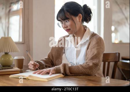A beautiful and focused young Asian woman in a cosy knitted sweater keeping her diary, writing something on a book, or doing homework at a table indoo Stock Photo