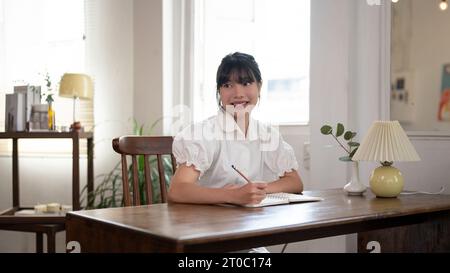 A beautiful and smiling young Asian woman keeping her diary or writing something on a book at a table in a minimal bright room. Stock Photo