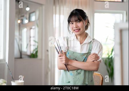 A beautiful and creative young Asian female artist in an apron smiles at the camera, stands with her arms crossed, and holds paint brushes in her hand Stock Photo