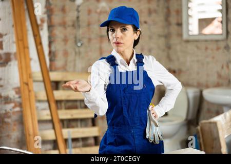 Female builder wearing overalls and cap posing at construction site, looking at camera Stock Photo