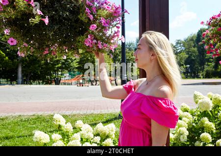 Pretty woman walks in the city park and holding Petunia flowers. She is smiling and wearing a pink off shoulder dress Stock Photo