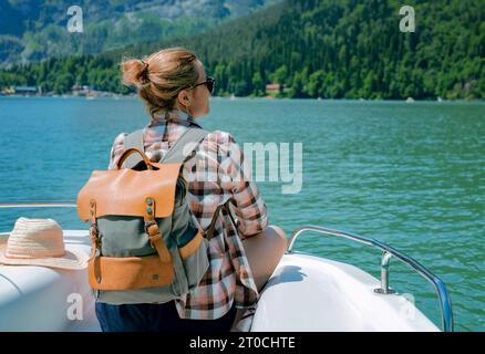 Hiking traveller woman with backpack enjoying beautiful view of turquois blue Lake Ritsa and mountains in Abkhazia sitting on deck of sailing paddlebo Stock Photo
