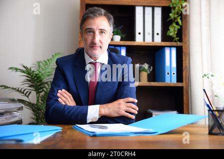 Businessman in his home office wearing blue suit and red tie with documents on wooden table and bookshelves background. Stock Photo