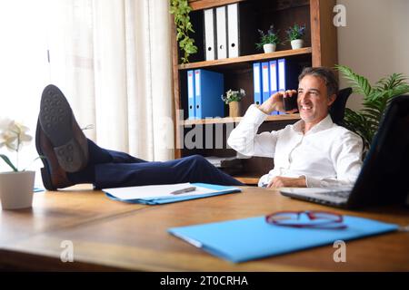 Smiling businessman talking on the mobile phone with feet on the table and documents and laptop on the table and shelves in the background in his offi Stock Photo