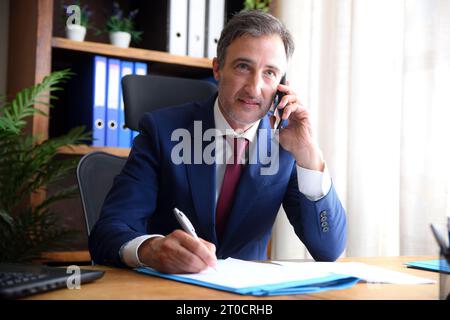 Businessman in blue suit and tie talking on mobile phone and writting in office with documents on the table and shelves in the background Stock Photo