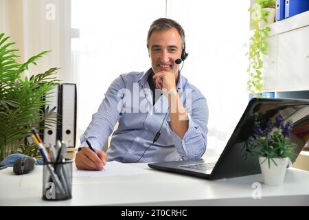 Smiling freelance in blue shirt doing video conference from a laptop listening with headphones and taking notes in an white office. Stock Photo
