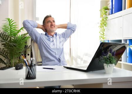 Imaginative entrepreneur freelancer in his home office sitting in a chair stretched out with his hands behind his head in a white office with shelves Stock Photo