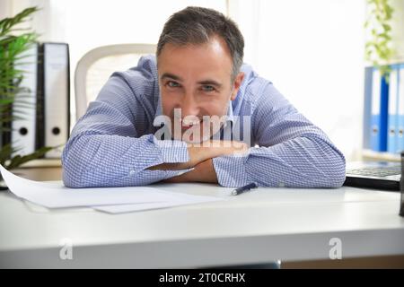 Freelance in blue shirt smiling and leaning on an white office table with papers and folders in the background Stock Photo