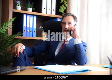 Elegant businessman in blue suit and red tie talking on mobile phone and pen in hand in office with documents on the table and shelves in the backgrou Stock Photo