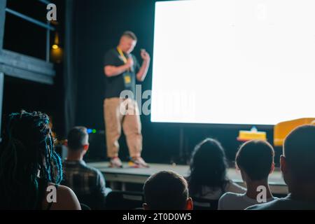 Male speaker standing on stage near big screen and giving presentation to participants sitting in dark hall during business seminar Stock Photo