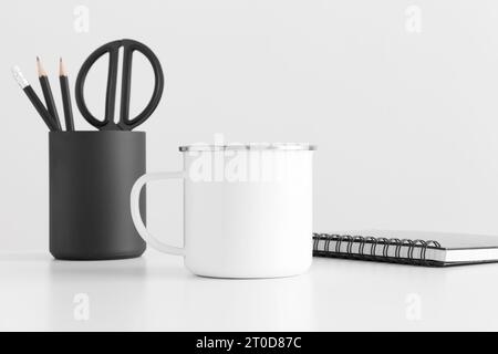 Enamel mug mockup with workspace accessories on a white table. Stock Photo