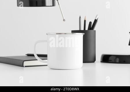 Enamel mug mockup with a lamp, notebook and workspace accessories on a white table. Stock Photo