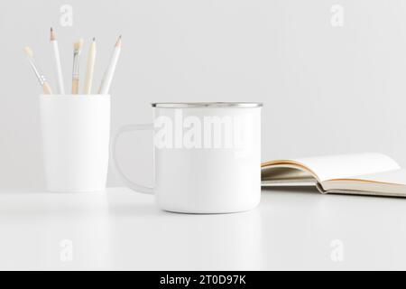 Enamel mug mockup with workspace accessories on a white table. Stock Photo