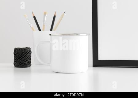 Enamel mug mockup with workspace accessories on a white table. Stock Photo