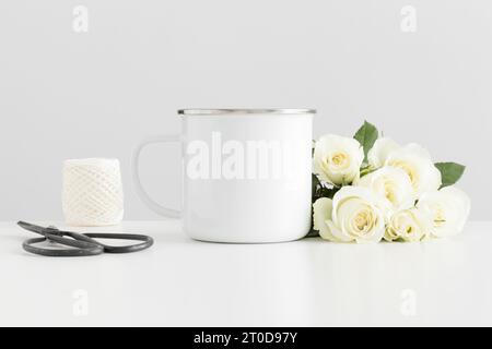 Enamel mug mockup with a bouquet of roses and workspace accessories on a white table. Stock Photo