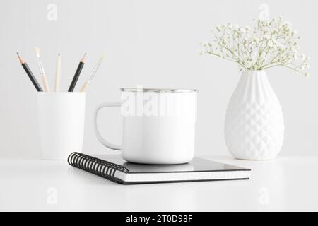 Enamel mug mockup with workspace accessories and a gypsophila on a white table. Stock Photo