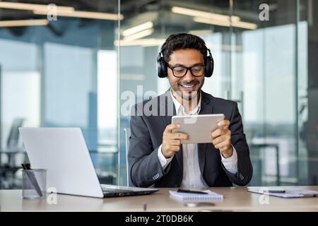 Businessman inside office with headphones and tablet computer watching online video stream, man in business suit smiling contentedly sitting at desk at workplace in business suit. Stock Photo