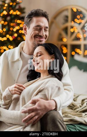 cozy living, happily married couple embracing each other near blurred Christmas lights on backdrop Stock Photo