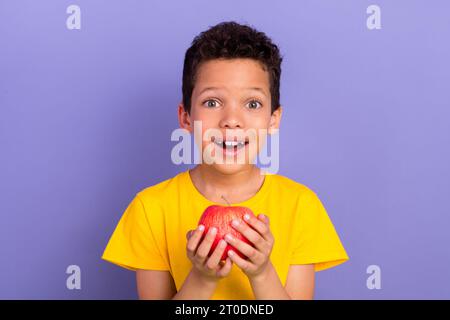 Photo portrait of charming young boy excited hold apple hungry wear yellow outfit isolated on purple color background Stock Photo