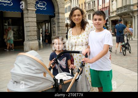 Happy multi-ethnic family, mother with children, boy and girl walking with baby pram, strolling the Italian medieval city during holidays. Family rela Stock Photo