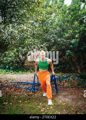 Non-binary man with green top and orange pants drags a garden hose behind him Stock Photo