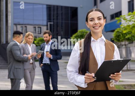 Portrait successful business woman from outside office building, female worker is satisfied smiling and looking at camera, holding papers documents in hands, working with colleagues and partners. Stock Photo