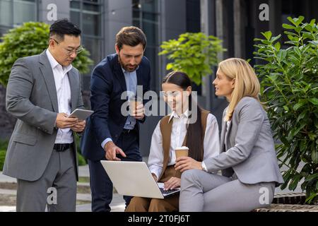 Four business people in business clothes outside office building happily chatting on lunch break, diverse team of men and women together, smiling and friendly communication. Stock Photo