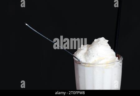 Milk shake on dark background. Close up of white vanilla milk shake with spoon and straw in tall glass mug. Restaurant, dinner or pub dessert backgrou Stock Photo