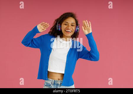 Pretty woman in headphones dancing with raised hands and looking at camera on pink studio background Stock Photo