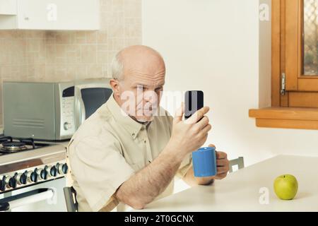 Struggling to see, a senior man finds difficulty with his smartphone while in his kitchen. Phones today may overlook those with eyesight issues. He co Stock Photo