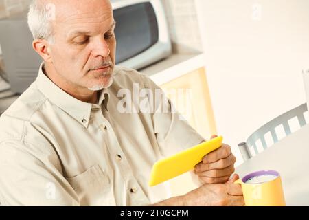 In his kitchen, a senior finds using his smartphone challenging. He squints over breakfast, indicative of phones not designed for vision issues. Perha Stock Photo