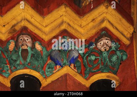 Bird pecks bunch of blue grapes, flanked by faces and green foliage, against a backdrop of vivid orange-red and yellow.  Sculpted capitals on left side of main portal of medieval Cathedral of Saint George in Limburg-an-der-Lahn, Hesse, Germany.  This Late Romanesque / Early Gothic cathedral, built in the late 1100s / early 1200s AD, was given back its original exuberant and colourful appearance through restoration work in the 1960s and ‘70s, with colours determined by traces of original paint. Stock Photo