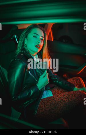 Urban photography with green and red neons in a car. Young blonde Caucasian model pose in a black leather jacket Stock Photo