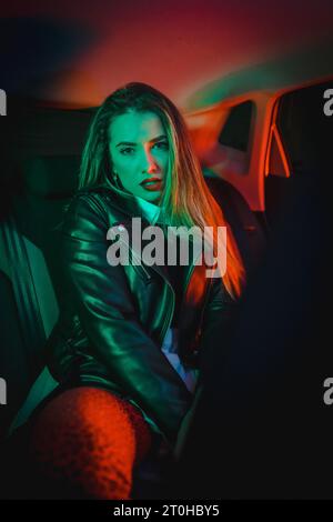 Urban photography with green and red neons in a car. Young blonde Caucasian model pose in a black leather jacket Stock Photo