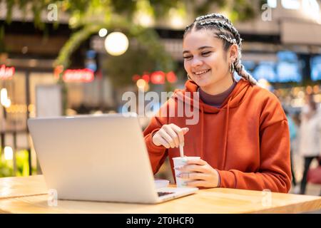 Alternative girl with white braids with a computer in a shopping center, making a video call with a hot coffee in her hands Stock Photo