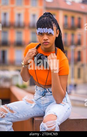Lifestyle with a young trap dancer with braids. Black-race girl pose with African ethnic group with orange shirt and cowboy pants sitting. With Stock Photo