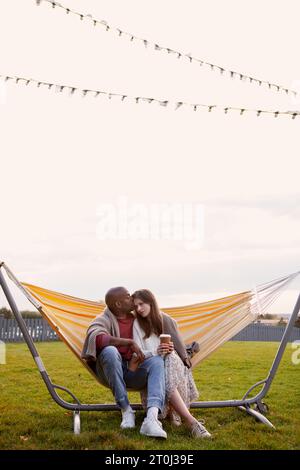 Romantic multiethnic couple with coffee in cup sitting comfortably in hammock in park, covered with cozy plaid, hugging. Stock Photo