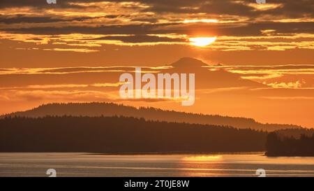 The sun as a large yellow ball above Mt Baker 130 km away is surrounded by clouds and an orange sky as seen over the water from Vancouver Island. Stock Photo