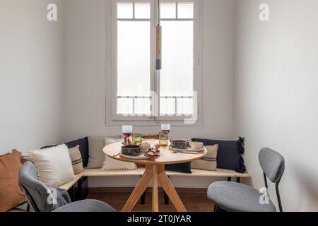 Horizontal shot breakfast for two with wine on a wooden round table gray chairs and a corner bench with pillows in front of large window in five-star Stock Photo